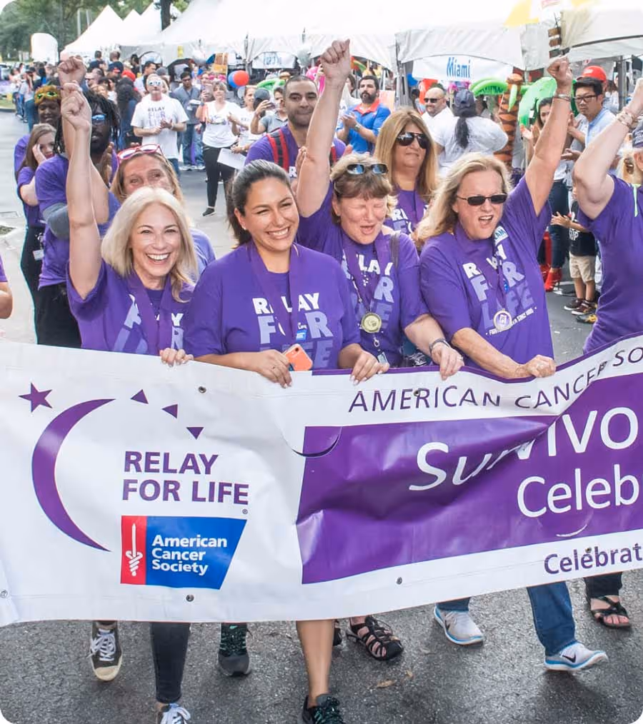 Group of smiling participants wearing purple 'Relay For Life' shirts, holding a banner for the American Cancer Society Relay For Life Survivors Celebration during a community event